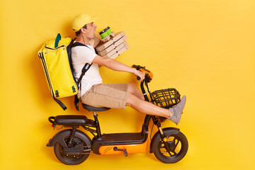 Side view of shocked scared man wearing white t shirt and cap riding electric scooter with backpack, holding pizza boxes and coffee in disposable cups, road accident, isolated over yellow background.