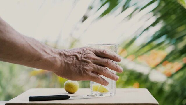 Close a glass of water , lime and knife on a wooden table.  Male hand takes a glass of water.In the background, palm leaves and the sea on a sunny summer day.