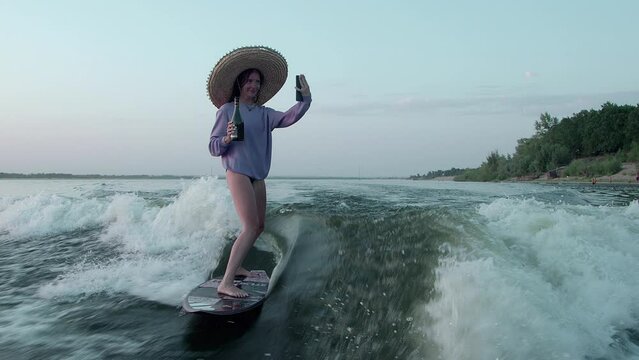 A female surfer in a straw hat jumps on a wakeboard with champagne in her hands, taking a selfie on her phone . An experienced wakeboarder sprays water drops into the camera.