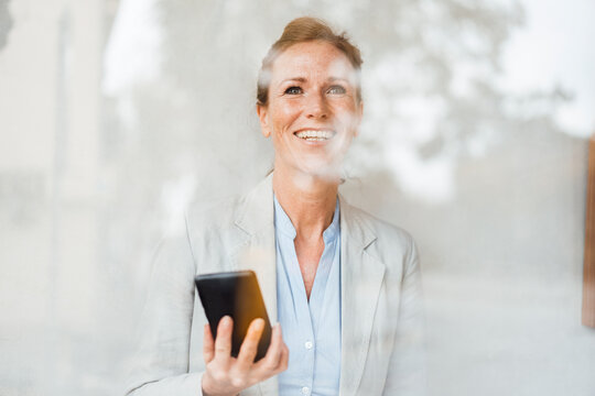 Happy Businesswoman Holding Mobile Phone In Cafe Seen Through Glass