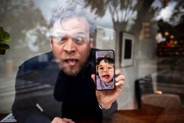 Businessman with eyes crossed showing daughter's face on video call in cafe