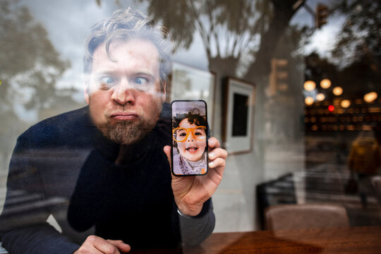 Playful businessman with eyes crossed showing daughter's face on video call in cafe