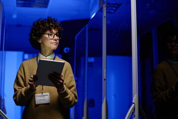 Young female engineer using tablet while walking in corridor of data center and checking hardware...