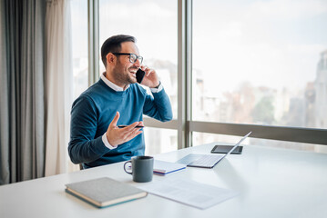Cheerful businessman talking on smart phone while sitting at office desk