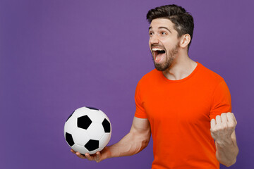 Young fun exultant happy fan man he 20s wearing orange t-shirt cheer up support football sport team hold in hand soccer ball watch tv live stream do winner gesture isolated on plain purple background.