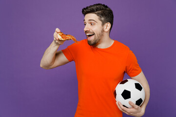 Young excited amazed fan man he wears orange t-shirt cheer up support football sport team hold in hand soccer ball eat slice of italian pizza watch tv live stream isolated on plain purple background.