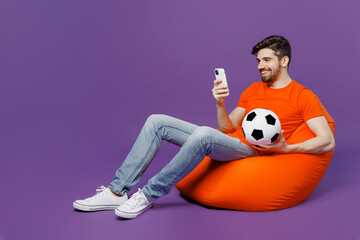 Full body young fan man he wear orange t-shirt cheer up support football sport team hold soccer ball mobile cell phone watch tv live stream sit in bag chair isolated on plain dark purple background.