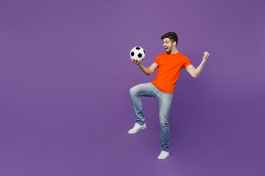 Full body young fan man he wear orange t-shirt cheer up support football sport team hold juggling on knee soccer ball watch tv live stream sit in bag chair isolated on plain dark purple background