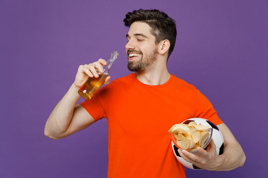 Young happy smiling satisfied fun fan man he in orange t-shirt cheer up support football sport team hold in hand soccer ball bottle drink beer watch tv live stream isolated on plain purple background.