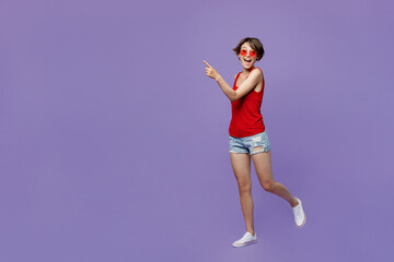 Fototapeta na wymiar Full body young smiling happy fun woman 20s she wear red tank shirt eyeglasses walking goign strolling point index finger aside on workspace area isolated on plain purple backround studio portrait