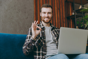 Young smiling fun man 20s he wears brown shirt hold use work on laptop pc computer show ok sitting on blue sofa in own living room apartment stay home indoor flat on weekends People lifestyle concept