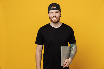 Young confident happy bearded tattooed man 20s he wear casual black t-shirt cap hold closed laptop...