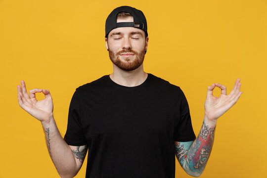 Young Bearded Tattooed Man 20s He Wears Casual Black T-shirt Cap Hold Spread Hands In Yoga Om Aum Gesture Relax Meditate Try To Calm Down Isolated On Plain Yellow Wall Background Tattoo Translate Fun