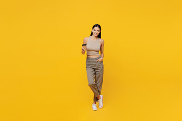 Full body young latin woman 30s she wear basic beige tank shirt hold takeaway delivery craft paper brown cup coffee to go isolated on plain yellow backround studio portrait. People lifestyle concept