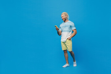 Full size side view young blond man with dreadlocks 20s he wear white t-shirt hold in hand use mobile cell phone isolated on plain pastel light blue background studio portrait People lifestyle concept