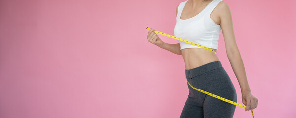 Slim woman in sportswear measures her waist using tape measure on pink background. diet woman and...