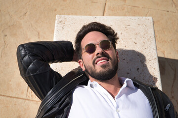Portrait of handsome young man with beard, sunglasses, leather jacket and white shirt, lying on a stone bench. Concept beauty, fashion, trend, peace, relax, tranquility.