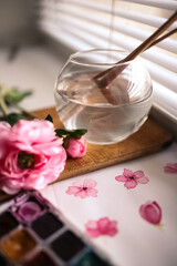 Round vase with water and brushes standing near the picture of pink flowers on the wood board and a lying pink flower