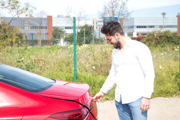 Handsome young man with beard, sunglasses, white shirt and jeans, opening the trunk of his red sports car. Concept beauty, fashion, trend, luxury, motor, sports, successful, luggage, trunk.