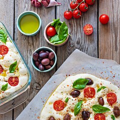 Fresh Italian flat bread (raw) Focaccia with tomatoes, olives, garlic and herbs on a wooden background.