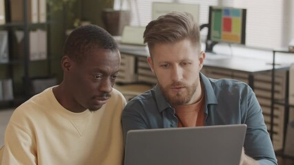 Chest up slowmo of two multiethnic male colleagues discussing ideas of new project while looking at laptop, working together in successful digital marketing agency