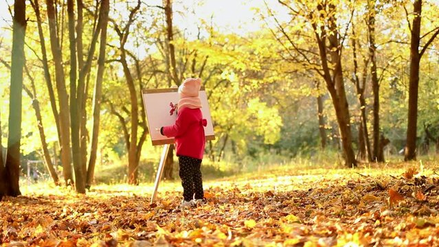 A little girl in a red coat paints an watercolor on easel in the park against the background of the autumn landscape. A cute child is taking paint from the palette, looks and smiles into the frame