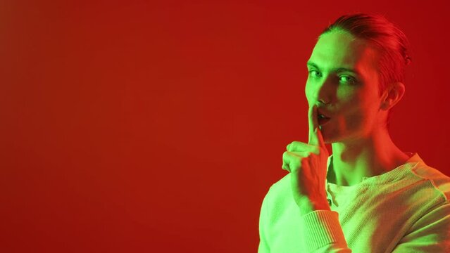 Man secret. Hush gesture. Intrigue conspiracy. Keep quiet. Green neon light smiling guy showing shh finger isolated on red color copy space advertising background.