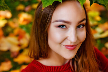 A large portrait of a young beautiful woman in a red sweater against the background of fallen yellow maple leaves. Autumn in the park.