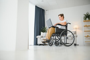 Middle age grey-haired disabled woman having video call sitting on wheelchair at home.