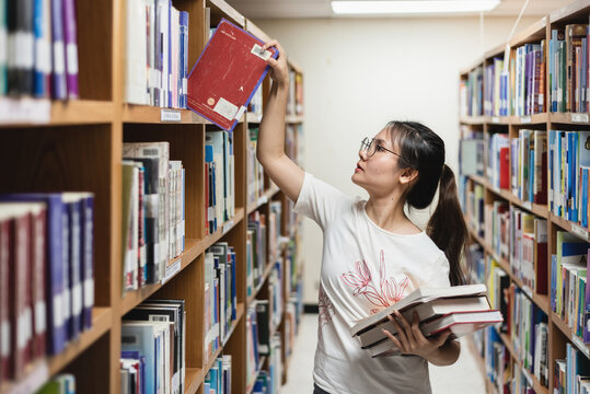 Concept library and learning, beautiful teenage Asian woman picking books from a bookshelf.