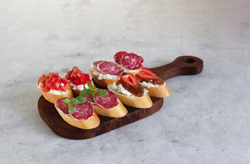 Bruschettas with salami, tomatoes, fig jam and strawberries on the brown wooden board. Gray background