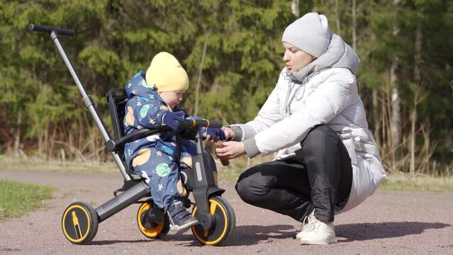 Mother with child on a walk in the spring park, kid on toddler tricycle push bike baby trike with parent handle.