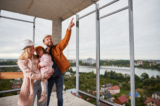 Woman Holding Daughter, Husband Pointing At Something At Construction Site. Family With Child Future Homeowners Observing Apartment Building Under Construction.