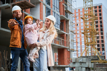 Fototapeta na wymiar Man, woman and child standing outside building under construction. Happy family homeowners posing on the street at construction site.