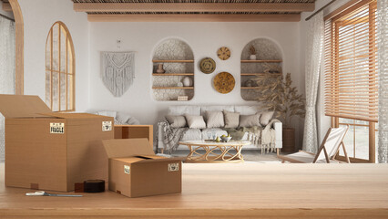Wooden table, desk or shelf with stack of cardboard boxes over blurred view of bsohemian living room in boho style, bohemian interior design, moving house concept with copy space
