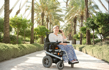 a smiling woman sitting in an electric wheelchair enjoys a sunny day in the garden of the city park...