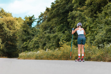Athletic young woman training on the roller ski at country road, back view. Biathlon ride with ski poles, in the helmet. Low angle view. Copy space. Concept of sports competition and summer workout