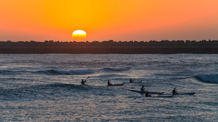 Athlete Paddlers Silhouetted Paddling Surf Ski Crafts Riding Ocean Waves at Sunrise with Harbor...