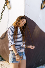 a girl with a dreadlocked hairstyle poses on the beach near a ship in summer, a sunny day, dressed in a T-shirt and denim shorts