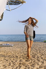 a girl with a dreadlocked hairstyle poses on the beach near a ship in summer, the sea and sand on a bright sunny day, dressed in a T-shirt and denim shorts