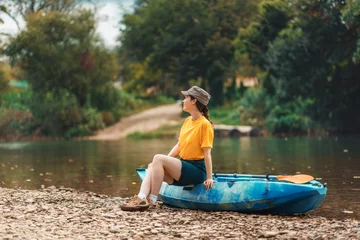 Foto op Canvas Summertime. A young smiling woman is sitting on a blue kayak and looking at the sky. The concept of kayaking and outdoor activities © _KUBE_