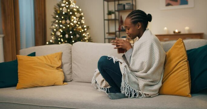 Winter evening. After hard day's work, young African-looking woman sat on sofa, covered herself with blanket, and drank hot, fragrant coffee. There are yellow pillows on the sofa. The girl relaxed.