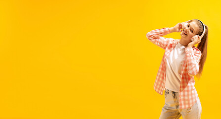 Portrait of young cheerful girl posing, listening to music in headphones isolated over yellow studio background. Flyer