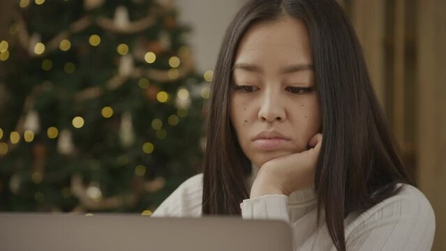 Young adult asian woman is bored in front of a laptop computer and Christmass tree in background