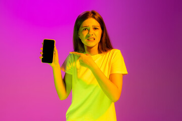 Portrait of young girl pointing at phone with displeased expression isolated over pink background in neon light