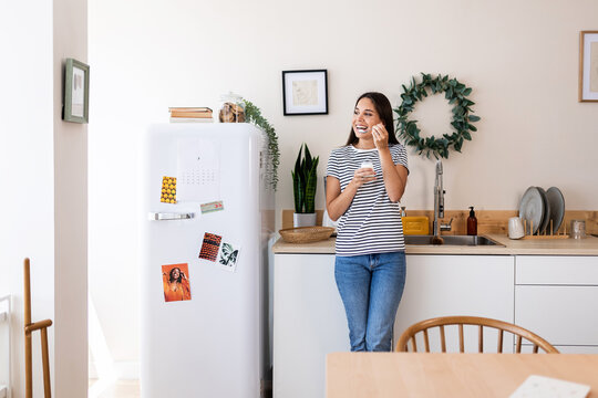 Pretty young adult woman eating natural yogurt while standing in the kitchen at home - Healthy lifestyle concept