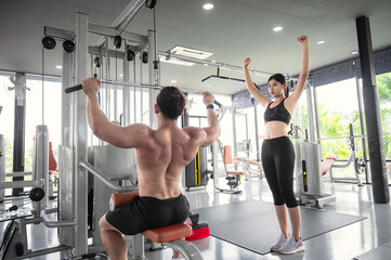 Fototapeta premium Young Asian bodybuilder trainer showing correct use of exercise equipment for Asian women to follow, in fitness room. Asian man trainer is teaching Asian women correct use of exercise equipment in gym