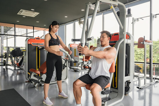 Young Asian bodybuilder trainer showing correct use of exercise equipment for Asian women to follow, in fitness room. Asian man trainer is teaching Asian women correct use of exercise equipment in gym