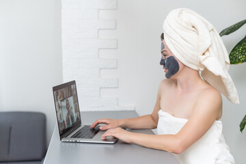 Young woman with towel on head and black facial clay mask sitting at home and using laptop computer . Staying at home, quarantine, distance learning, online education, technology, modern lifestyle