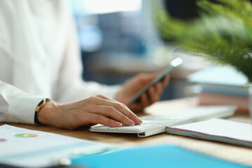 Female hands hold smartphone and work on laptop keyboard